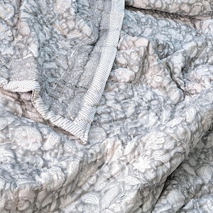 May include: A gray and white floral patterned quilt with a quilted design. The quilt features a detailed floral pattern and a striped border. The fabric is soft and appears to be cotton or a similar material. The quilt is folded and draped.