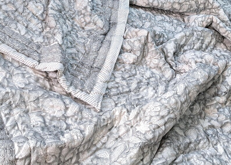 May include: A gray and white floral patterned quilt with a quilted design. The quilt features a detailed floral pattern and a striped border. The fabric is soft and appears to be cotton or a similar material. The quilt is folded and draped.