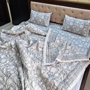 May include: A gray and white floral patterned quilt set on a bed. The set includes a quilt and two matching pillowcases. The bed has a beige upholstered headboard and a dark brown wooden frame. The quilt is draped over the bed.