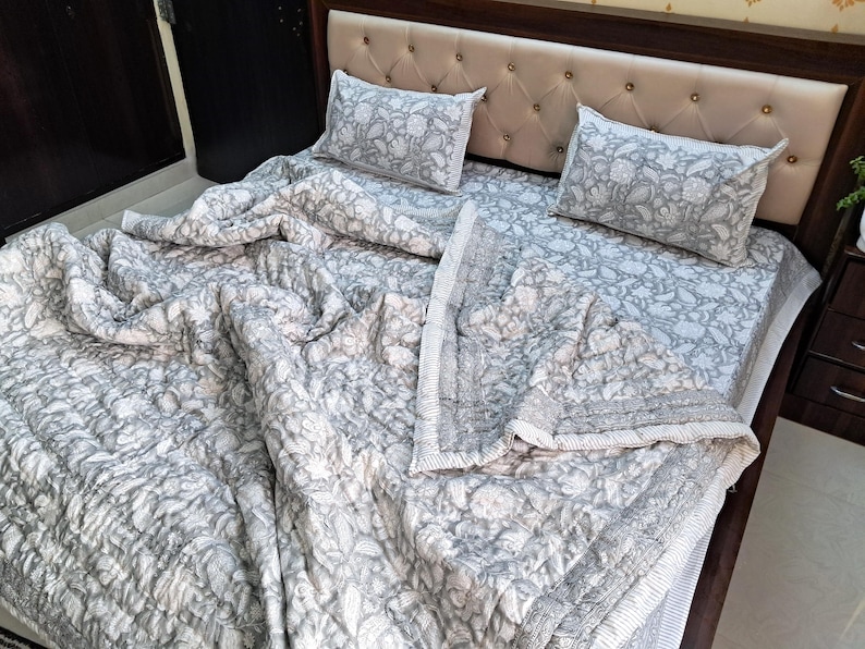 May include: A gray and white floral patterned quilt set on a bed. The set includes a quilt and two matching pillowcases. The bed has a beige upholstered headboard and a dark brown wooden frame. The quilt is draped over the bed.