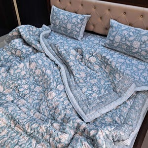 May include: A blue floral patterned quilt set with two matching pillowcases on a bed. The quilt and pillowcases feature a repeating pattern of white and peach-colored flowers and leaves. The bedspread has a striped border.