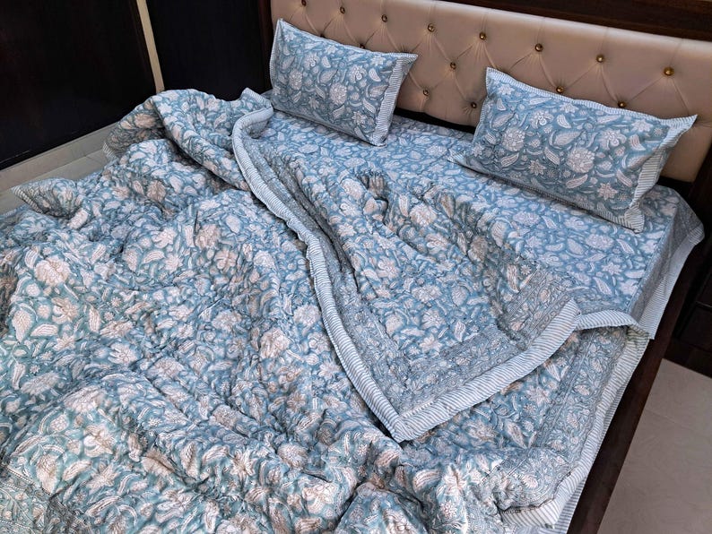 May include: A blue floral patterned quilt set with two matching pillowcases on a bed. The quilt and pillowcases feature a repeating pattern of white and peach-colored flowers and leaves. The bedspread has a striped border.
