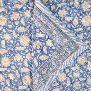May include: A blue and white floral patterned quilt. The quilt features a repeating pattern of beige flowers and leaves on a blue background. The border has a striped design with floral accents.