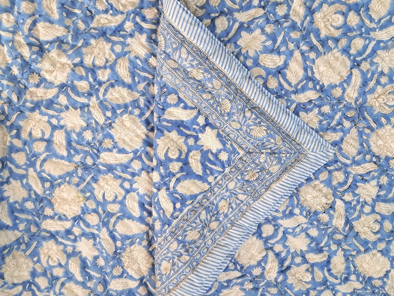 May include: A blue and white floral patterned quilt. The quilt features a repeating pattern of beige flowers and leaves on a blue background. The border has a striped design with floral accents.
