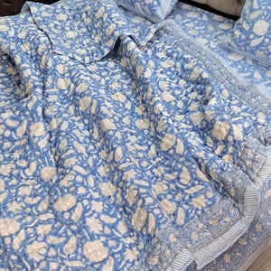May include: A blue and white floral patterned quilt set. The bedding features a repeating pattern of flowers and leaves in white against a light blue background. The set includes a quilt and pillowcases.