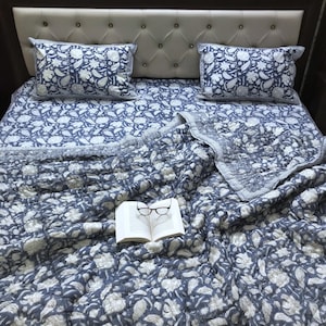 May include: A blue and white floral patterned bedding set, including a quilt and two pillowcases, rests on a bed with a tufted headboard. An open book with eyeglasses is on the quilt. The bedding has a floral design.