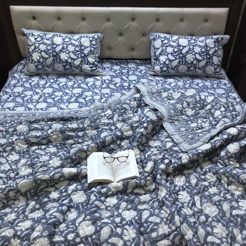 May include: A blue and white floral patterned bedding set, including a quilt and two pillowcases, rests on a bed with a tufted headboard. An open book with eyeglasses is on the quilt. The bedding has a floral design.