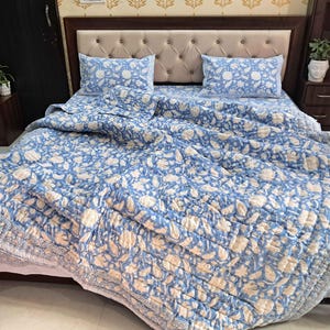 May include: A blue and white floral patterned quilt set on a bed, including a quilt, two pillowcases, and a fitted sheet. The bedding has a quilted design and a light blue and white color scheme. The bed has a beige headboard.