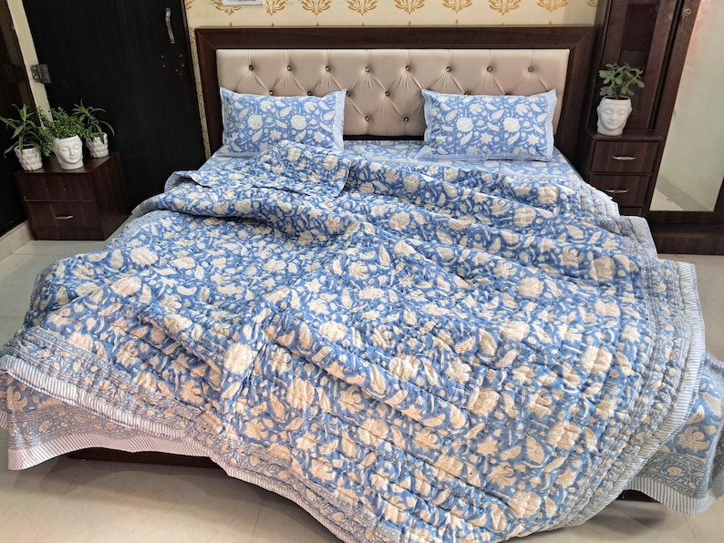 May include: A blue and white floral patterned quilt set on a bed, including a quilt, two pillowcases, and a fitted sheet. The bedding has a quilted design and a light blue and white color scheme. The bed has a beige headboard.