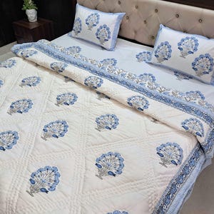 May include: A white bedspread and pillow set with a blue floral pattern. The bedding features a quilted design with a repeating motif of stylized blue flowers. The pillows and the border of the bedspread have a blue striped trim.