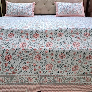 Puede incluir: Juego de cama blanco con estampado floral, con flores rojas y hojas verdes. Se ven fundas de almohada a juego con borde rosa. La colcha tiene un borde a rayas. El patrón floral se repite.
