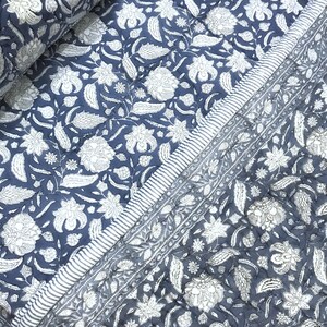 May include: A quilted textile with a floral pattern in white against a navy blue background. The design features stylized flowers and leaves. A striped border in white and navy blue frames the edge of the textile.