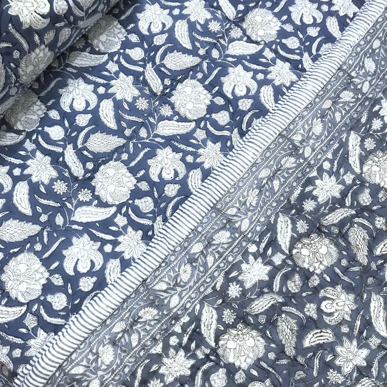 May include: A quilted textile with a floral pattern in white against a navy blue background. The design features stylized flowers and leaves. A striped border in white and navy blue frames the edge of the textile.