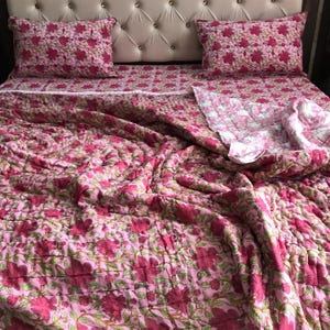 Op de afbeelding: Een roze quilt met bloemenpatroon, een wit laken met bloemenpatroon en twee roze kussenslopen met bloemenpatroon. De quilt is gemaakt van katoen en is perfect voor een slaapkamer.