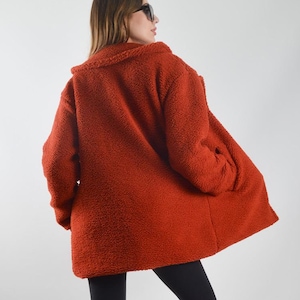 Op de afbeelding: Een vrouw draagt een lange, roestkleurige, pluche jas met een zwarte legging en zwarte laarzen.