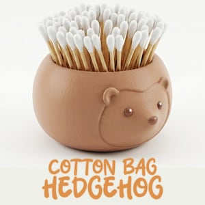 Può includere: Un contenitore marrone a forma di riccio pieno di bastoncini di cotone con punte bianche. Il contenitore ha una superficie strutturata e un simpatico design del viso. Il testo "COTTON BAG HEDGEHOG" è visualizzato in lettere arancioni nella parte inferiore dell'immagine.