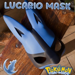 May include: A blue and black Lucario mask with the text "LUCARIO MASK" in a bubble font. The mask is fan-made and features the Pokemon logo. The mask is on a wooden surface.