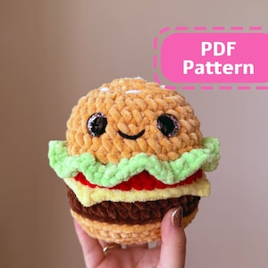 Op de afbeelding: Een gehaakt pluche speelgoed in de vorm van een hamburger met een glimlachend gezicht. Het speelgoed is gemaakt van bruin, geel, groen en rood garen. Het speelgoed is verkrijgbaar met een PDF-patroon.