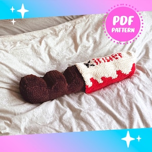 May include: A crocheted chocolate bar cushion in brown, red, and white, designed to look like a Kinder chocolate bar. The word "Kinder" is displayed in red. The image also includes a pink circle with the text "PDF PATTERN".