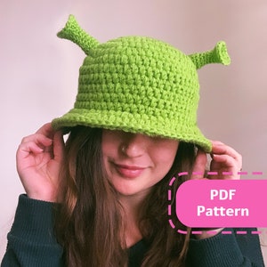 Puede incluir: Sombrero de crochet verde con dos cuernos en forma de orejas, que se asemeja a un personaje de la película Shrek. El sombrero lo lleva puesto una persona, y el texto "PDF Pattern" es visible en la imagen.