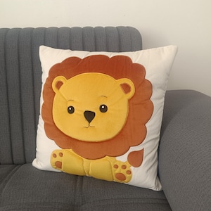 Puede incluir: Un cojín cuadrado decorativo con un diseño de león. El león tiene una cara y patas amarillas, una melena marrón y está sobre un fondo blanco. El cojín está en un sofá gris.
