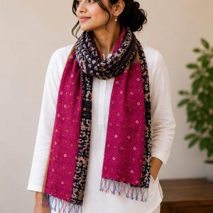 May include: A magenta scarf with a patterned design and fringed edges. The scarf features a dark blue and tan border with a repeating pattern. The scarf is draped over a white shirt.