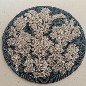 May include: A circular beaded placemat featuring a dark blue backdrop and a coral reef design in cream and beige tones. The coral is meticulously detailed, giving a textured look. This placemat is a decorative element for the dining table.