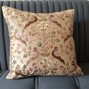 Può includere: Cuscino quadrato decorativo con un disegno ricamato di uccelli marroni appollaiati su rami con fiori. Il cuscino ha uno sfondo beige ed è posto su una superficie grigia.