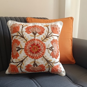 May include: A square throw pillow featuring an off-white fabric base with intricate, raised embroidery in shades of orange, peach, and brown, depicting a central floral mandala pattern. Behind it sits a solid, plain orange square throw pillow.
