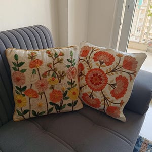 Peut inclure: Deux coussins décoratifs brodés avec des motifs floraux. L'un présente une variété de fleurs colorées dans des tons de rose, d'orange et de jaune. L'autre coussin a un grand motif floral circulaire dans des tons d'orange et de rose. Les deux coussins ont un fond crème et sont posés sur un canapé gris.