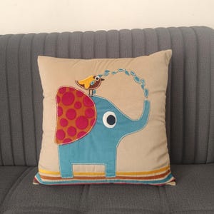 May include: Beige square decorative pillow featuring a whimsical elephant design. The blue elephant has a red and orange polka-dotted blanket and a small bird on its back. The pillow has a striped border with yellow, brown, and blue accents.