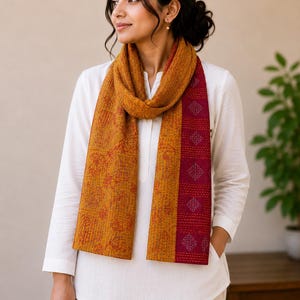 May include: Mustard yellow and burgundy scarf with a geometric pattern. The scarf is draped over a white long-sleeved shirt. The scarf has a textured appearance and a stitched design.