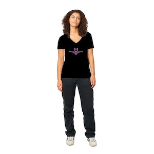 Könnte beinhalten: Schwarzes V-Ausschnitt-T-Shirt mit einem rosa Grafikdesign auf der Vorderseite. Die Grafik zeigt eine stilisierte Fledermaus oder ein ähnliches Wesen. Die Person trägt eine schwarze Hose und weiße Turnschuhe.