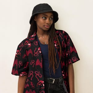 May include: A black button-up shirt with a red and black skull and snake print. The shirt has short sleeves and a collared neckline. The model is wearing a black bucket hat and has long brown braids.