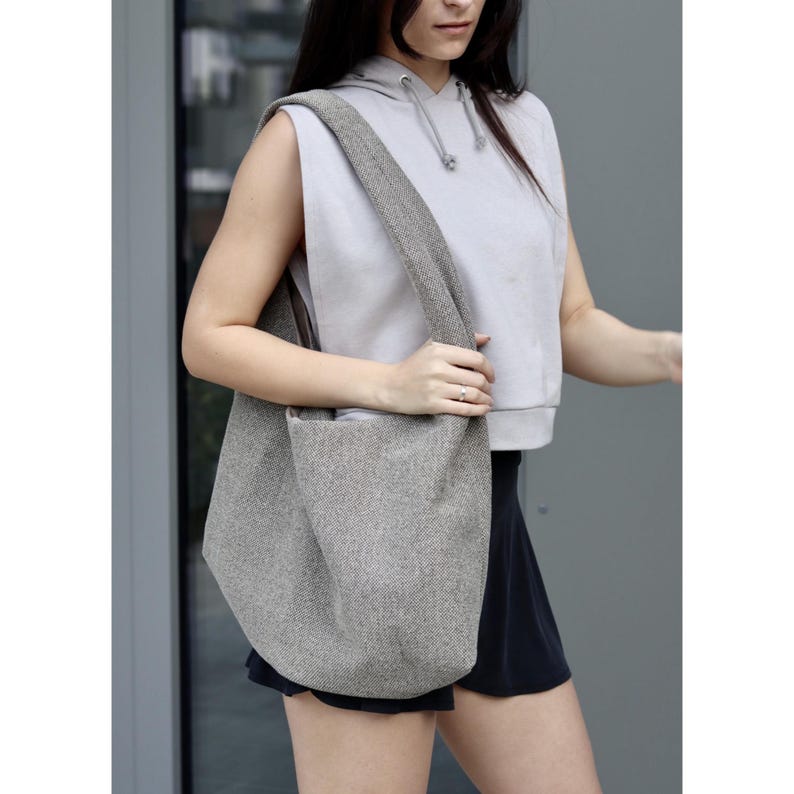 Pu&ograve; includere: Una borsa tote di maglia grigia con un design morbido. La borsa &egrave; indossata sulla spalla di una persona che indossa una felpa grigia senza maniche e pantaloncini neri.