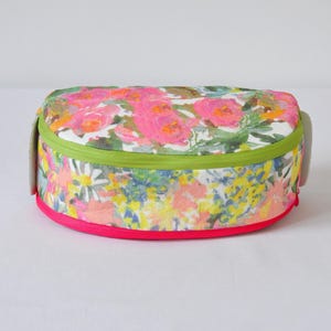May include: A floral print oval-shaped zippered pouch with a green zipper and a pink trim. The pouch is made of a soft fabric and has a white background with pink, yellow, and green flowers.