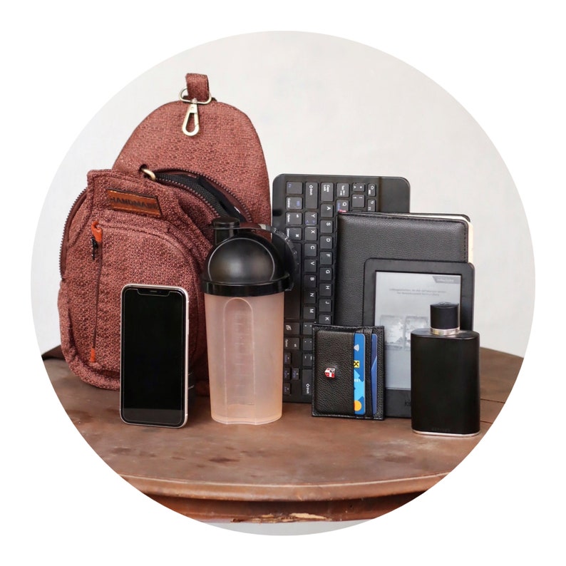 May include: A brown, textured, sling bag with a zippered pocket, a black and white keyboard, a black e-reader, a black wallet with credit cards, a clear plastic shaker bottle with a black lid, and a black bottle of perfume on a wooden table.