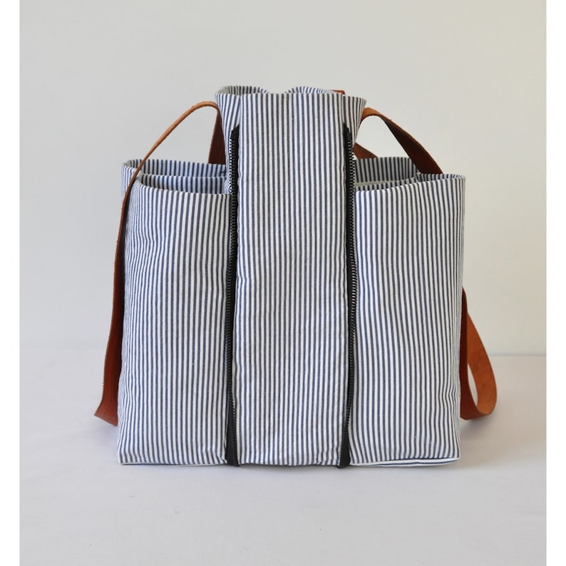 Puede incluir: Una bolsa de lona rayada azul y blanca con correas de cuero marr&oacute;n y cierres de cremallera negros.