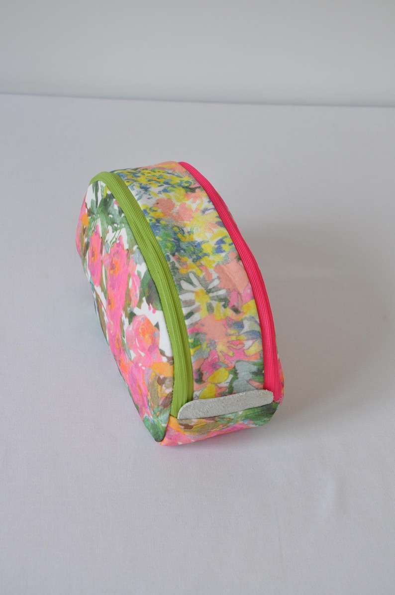 May include: A floral print makeup bag with a green and pink zipper. The bag is oval shaped and has a white fabric lining.