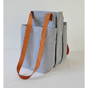 Puede incluir: Una bolsa de lona rayada azul y blanca con correas de cuero marr&oacute;n. La bolsa tiene varios bolsillos y un cierre de cremallera.