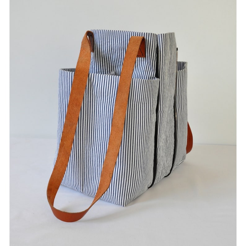 Puede incluir: Una bolsa de lona rayada azul y blanca con correas de cuero marr&oacute;n. La bolsa tiene varios bolsillos y un cierre de cremallera.