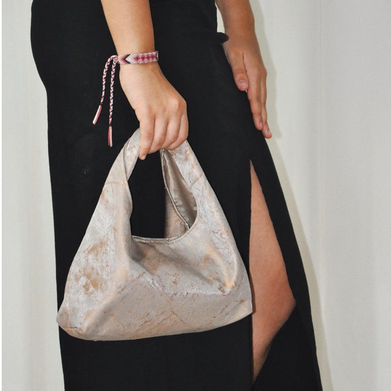 May include: A light beige handbag with an unusual shape, held by a person wearing a pink and white bracelet. The bag has a textured surface with brown and beige tones. The person is wearing a black dress.