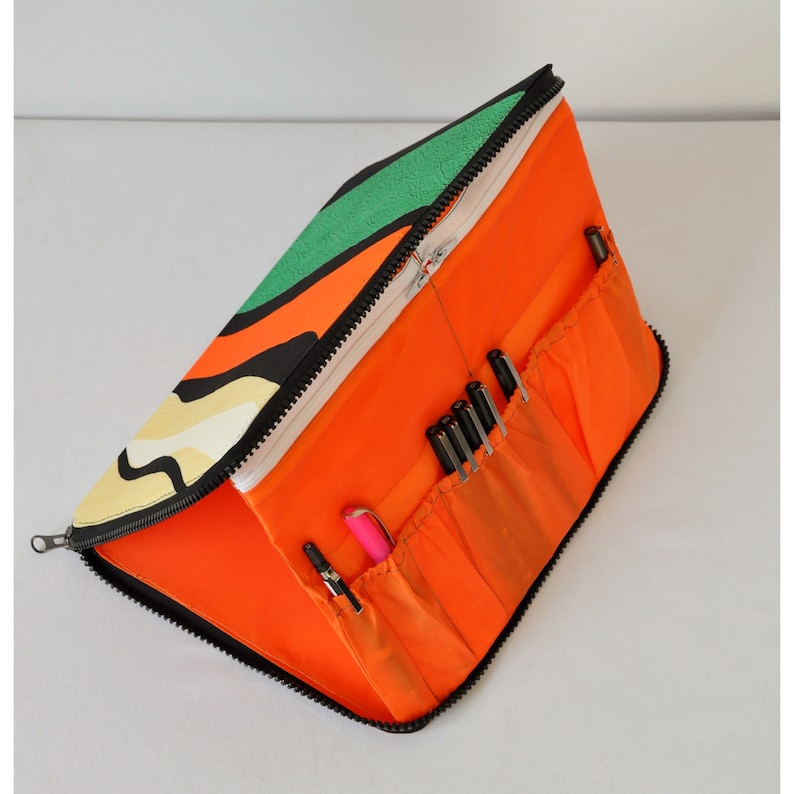 May include: A bright orange zippered pencil case with multiple pockets and a colorful abstract design on the exterior. The case is open and filled with black and pink pens.