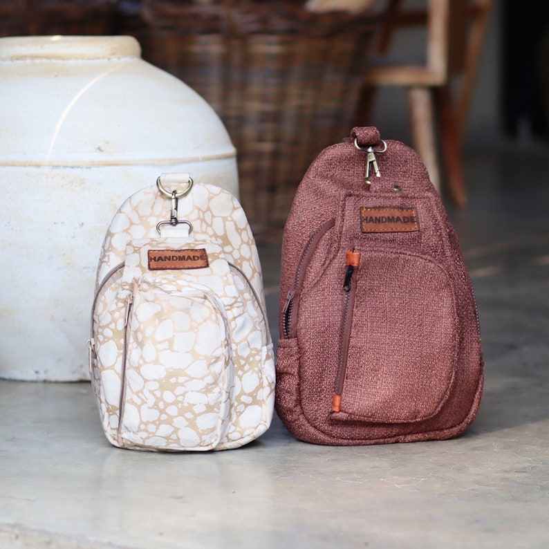 May include: Two handmade crossbody bags. One bag is white with a brown speckled pattern and the other is brown. Both bags have a brown leather tag that says "HANDMADE".