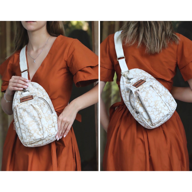May include: A white crossbody bag with a gold pattern and a brown leather tag that says "Handmade". The bag has a zipped pocket on the front and an adjustable strap.