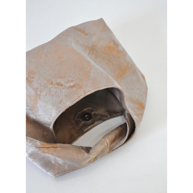 May include: A beige tote bag with a soft, velvety texture and a subtle, marbled pattern in shades of gold. The bag features a single handle and a magnetic snap closure. The interior is a darker brown colour.