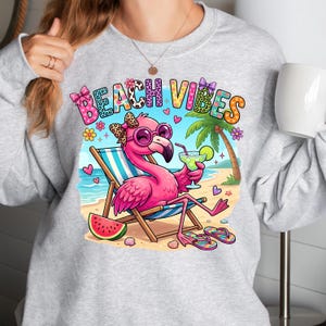 May include: A light gray sweatshirt featuring a colorful graphic of a pink flamingo wearing sunglasses and a bow, relaxing on a beach chair with a drink. The words "BEACH VIBES" are above the flamingo. A slice of watermelon and flip-flops are also visible.