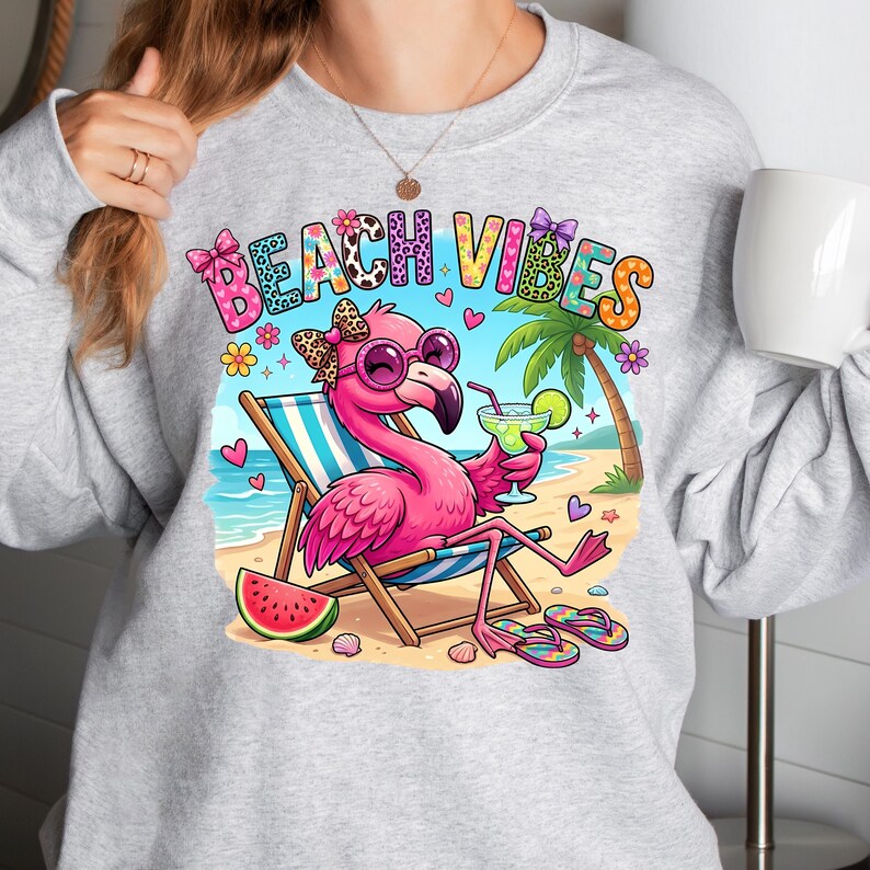 May include: A light gray sweatshirt featuring a colorful graphic of a pink flamingo wearing sunglasses and a bow, relaxing on a beach chair with a drink. The words "BEACH VIBES" are above the flamingo. A slice of watermelon and flip-flops are also visible.