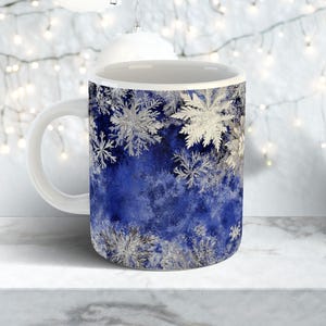 May include: White ceramic mug with a blue background and silver snowflake design. The snowflakes vary in size and are clustered around the top and bottom of the mug. The mug has a white handle and a white interior.