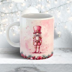 May include: A white ceramic mug featuring a pink watercolor design of a nutcracker soldier in a tall hat and uniform. The mug has a white handle and a decorative border of festive elements at the base, perfect for holiday beverages.